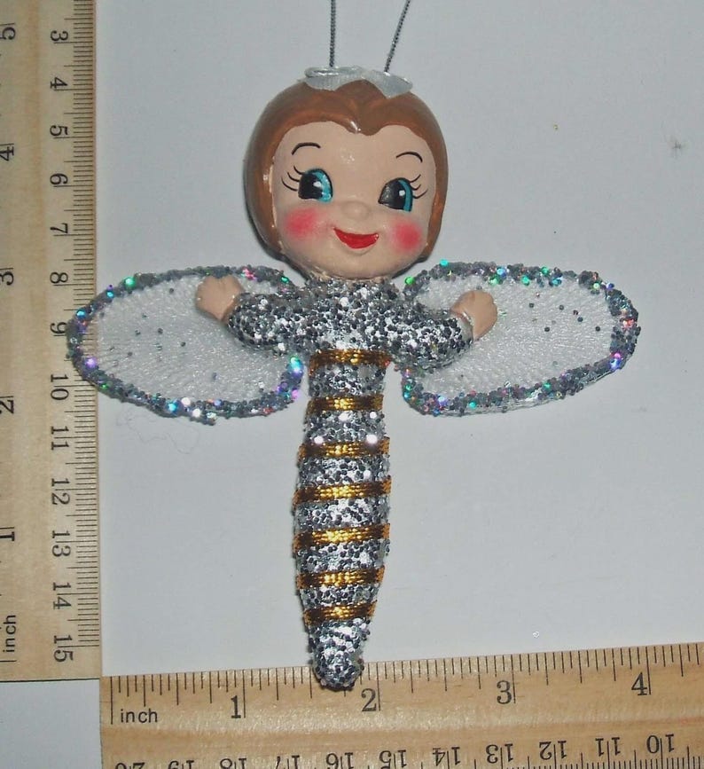 May include: A bee-themed ornament with a doll-like face, brown hair, and blue eyes. The body is covered in silver glitter with gold stripes and white glittery wings. A ruler shows the ornament is approximately 11.5 cm tall and 11.5 cm wide.