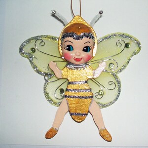 May include: A hanging ornament shaped like a bee character with a painted face, rosy cheeks, and silver glitter hair. The figure wears a yellow and silver striped outfit with sheer, pale yellow wings featuring silver glitter edges and swirls.