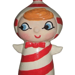 May include: A ceramic figurine of a child-like figure with a red bow tie and red and white striped clothing. The figure has orange hair, blue eyes, and a smiling expression. The head covering is white with red polka dots.