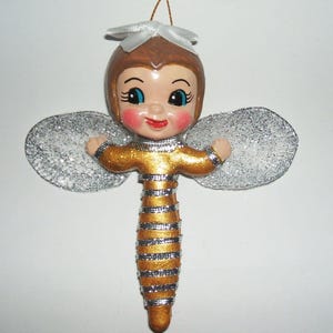 May include: A hanging ornament shaped like a bee with a human-like face, brown hair, and a white ribbon bow. The body features gold and silver horizontal stripes, and the wings are large, oval-shaped, and covered in silver glitter.