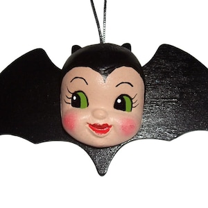May include: A hanging ornament featuring a black bat silhouette with a central doll-like face. The face has pale skin, bright green eyes, long black eyelashes, rosy pink cheeks, and red lips. A thin silver string is attached to the top for hanging.