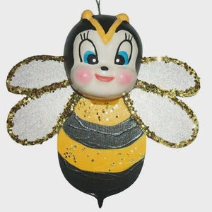 May include: A whimsical bee ornament with a smiling face, rosy cheeks, and blue eyes. The bee's body is yellow and black with glitter accents on the wings and body. A decorative item for a nursery or a child's room.