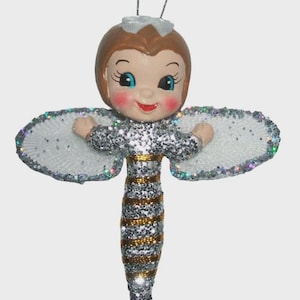 May include: A hanging ornament shaped like a bee with a human face. The figure has brown hair, blue eyes, and rosy cheeks. Its body is covered in silver glitter with horizontal gold stripes. It features white, glittery, oval-shaped wings.