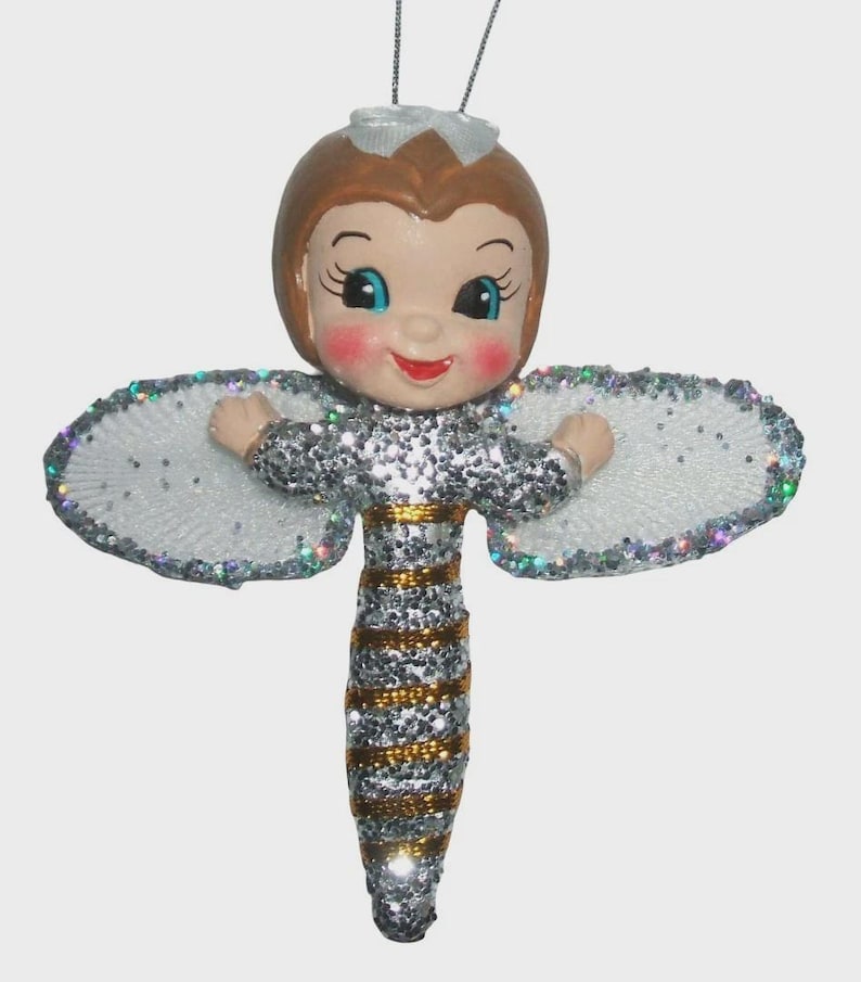 May include: A hanging ornament shaped like a bee with a human face. The figure has brown hair, blue eyes, and rosy cheeks. Its body is covered in silver glitter with horizontal gold stripes. It features white, glittery, oval-shaped wings.