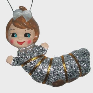 May include: A hanging ornament shaped like a caterpillar with a human-like face, featuring brown hair with a white bow, blue eyes, and rosy cheeks. The body is covered in silver glitter with gold ribbon accents wrapped around the segmented form.