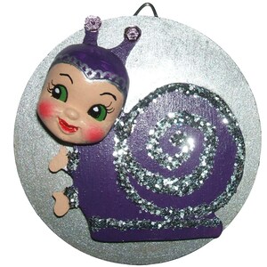 May include: A whimsical, purple snail ornament with a smiling face, green eyes, and glitter accents. The snail's shell features a silver glitter spiral design. The ornament is attached to a silver circular background.