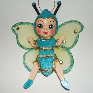 May include: A hanging ornament shaped like a fairy with a blue glittery outfit, gold trim, and pale yellow wings with green glitter edges. The figure has a blue cap with black antennae, pink cheeks, and blue shoes, suspended by a thin gold cord.