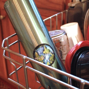 May include: A stainless steel water bottle with a blue and green earth sticker that says "Our Earth, Our Home". The bottle is in a dishwasher rack.