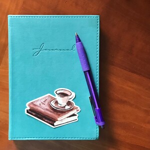 May include: A teal blue journal with the word "Journal" embossed on the front cover. A purple pen and a sticker of a cup of coffee on a stack of books are on the cover.