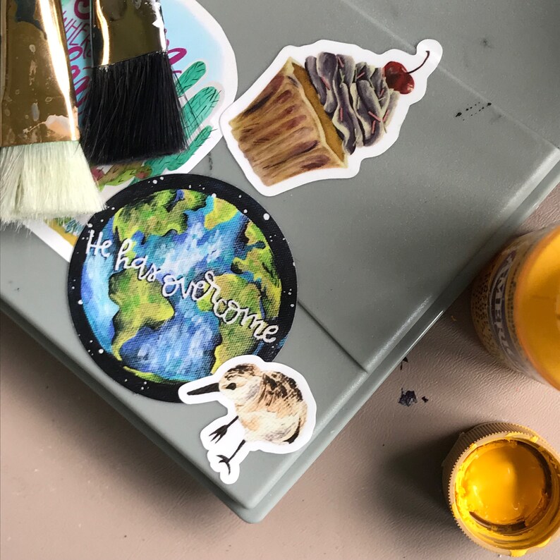 May include: A set of four stickers featuring a cupcake with a cherry on top, a bird, a globe with the text "He has overcome", and a cactus. The stickers are on a gray surface.