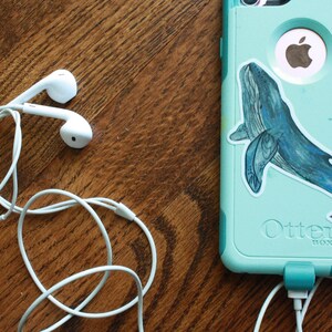 May include: White wired earphones with a tangle of cord on a wooden surface. A teal phone case with a whale sticker and the text "Otter Box" is visible in the background.