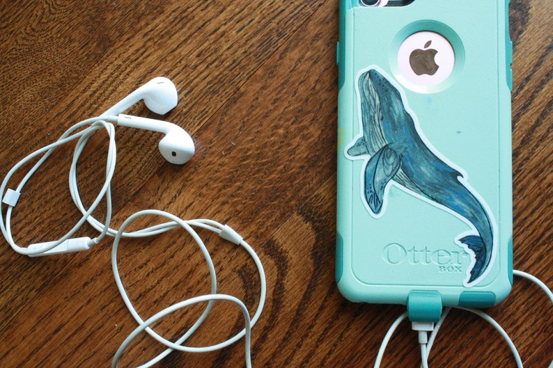 May include: White wired earphones with a tangle of cord on a wooden surface. A teal phone case with a whale sticker and the text "Otter Box" is visible in the background.