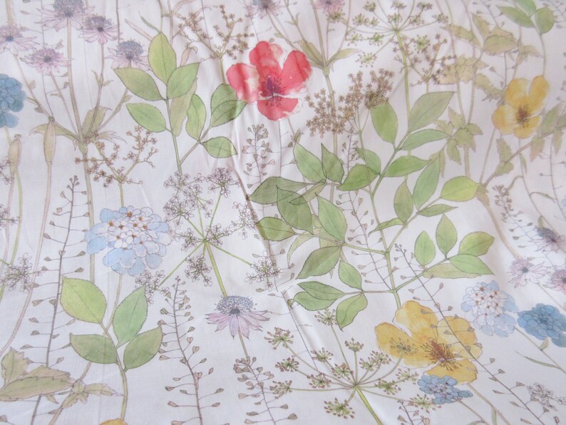 May include: A white fabric with a floral pattern. The pattern features various flowers, leaves, and stems in shades of green, blue, yellow, and red. The background is a light cream color.
