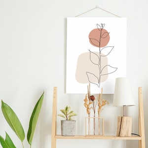 May include: A wooden shelf with a white wall in the background. The shelf has a lamp with a white shade, a potted plant, a vase with dried flowers, and a framed print with a line drawing of a plant with brown and beige shapes.