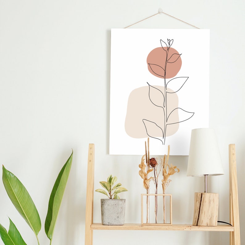 May include: A wooden shelf with a white wall in the background. The shelf has a lamp with a white shade, a potted plant, a vase with dried flowers, and a framed print with a line drawing of a plant with brown and beige shapes.