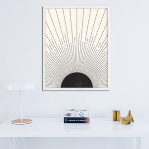 May include: A framed art print featuring a minimalist design of a black semi-circle with radiating lines against a cream background. The artwork is in a white frame. A white desk lamp and two books are in the foreground.