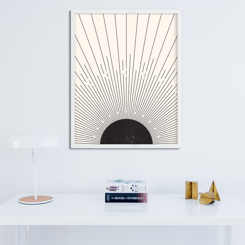 May include: A framed art print featuring a minimalist design of a black semi-circle with radiating lines against a cream background. The artwork is in a white frame. A white desk lamp and two books are in the foreground.