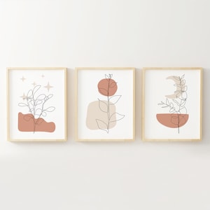 May include: Set of three minimalist line art prints featuring abstract shapes and foliage in shades of brown, beige, and white. The prints are framed in natural wood.
