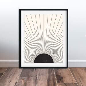 May include: Framed art print featuring a minimalist sunburst design. The artwork has a black semi-circle at the bottom, with black lines radiating upwards against a cream-colored background. The print is encased in a black frame, suitable for wall decor.