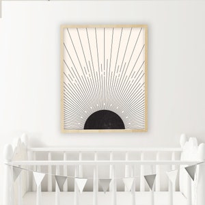 May include: A framed art print featuring a black and cream sunburst design. The artwork has a black semi-circle at the bottom with radiating lines extending upwards. The frame is a light wood color. The print is displayed above a white crib.