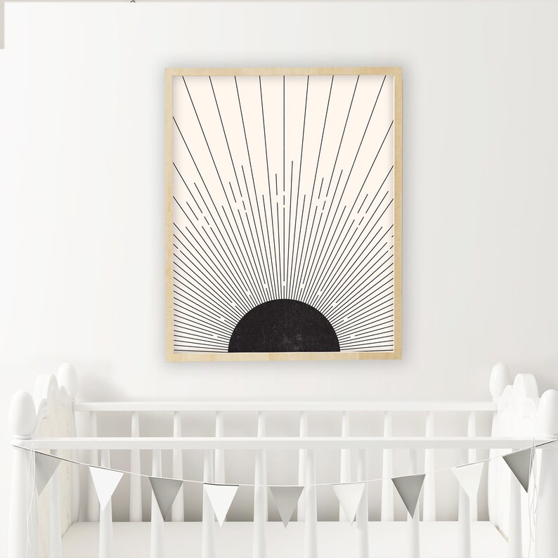 May include: A framed art print featuring a black and cream sunburst design. The artwork has a black semi-circle at the bottom with radiating lines extending upwards. The frame is a light wood color. The print is displayed above a white crib.