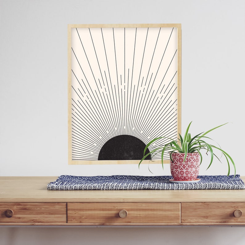 May include: A framed art print featuring a black and white sunburst design. The artwork is in a light wood frame and is displayed on a wooden table. A small potted plant with green leaves sits on the table, adding a touch of nature.
