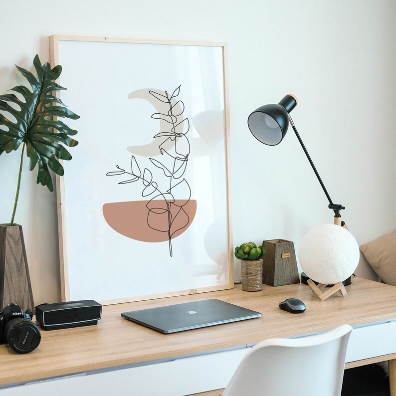 May include: A framed print of a minimalist line drawing of a plant with a peach colored background. The print is on a desk with a laptop, a lamp, and other decor.
