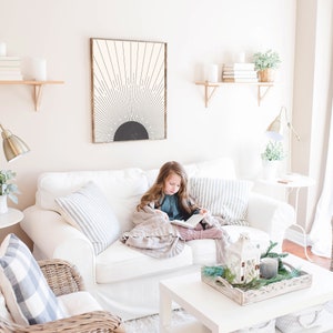 May include: A living room scene featuring a white sofa with striped and solid pillows. A framed abstract art piece hangs above the sofa. A young person is reading a book while wrapped in a blanket. Decorative items include candles and plants.