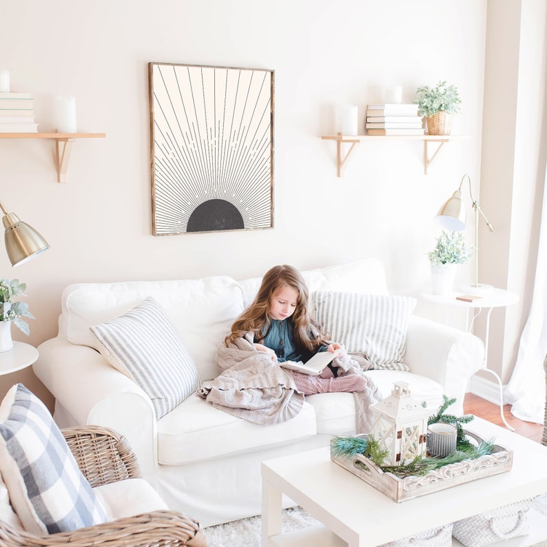 May include: A living room scene featuring a white sofa with striped and solid pillows. A framed abstract art piece hangs above the sofa. A young person is reading a book while wrapped in a blanket. Decorative items include candles and plants.