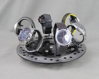 May include: A circular watch display stand made from a silver-colored disc, holding six wristwatches. The watches have various band colors, including black, brown, and yellow. The stand is on a neutral gray background.