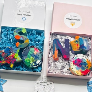 May include: Two gift boxes with Hanukkah-themed crayons. One box contains an "S" crayon, a Star of David, and a menorah-shaped crayon. The other box has an "N" crayon, a dreidel, and a "Happy Hanukkah" crayon. Each box has a sticker with a name and "Happy Hanukkah!"