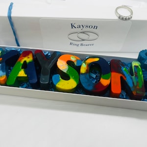 May include: A white box with the name Kayson and the words Ring Bearer printed on the lid. The box contains colorful crayon letters spelling out the name Kayson, blue shredded paper, and a blue crayon ring and dinosaur.