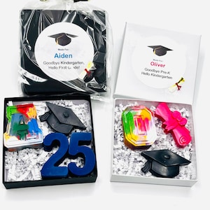 May include: Graduation-themed gift sets. One set is in a black box with a clear wrap and gold ribbon, labeled "Aiden." The other is in a white box labeled "Oliver." Each set includes a graduation cap and a diploma-shaped crayon. Additional sets include number and letter crayons.
