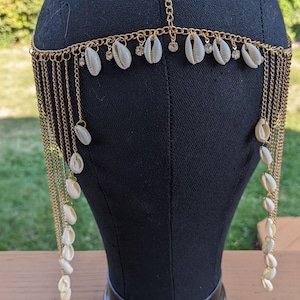 May include: Gold-toned headpiece featuring a chain design with dangling strands. Adorned with small white seashells and clear rhinestones. The headpiece is displayed on a black mannequin head, showcasing its intricate details.