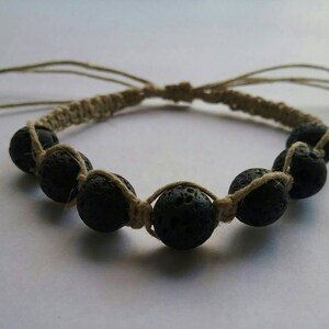 Puede incluir: Un brazalete de piedra de lava negra con cordón beige. El brazalete tiene un diseño anudado y es ajustable.