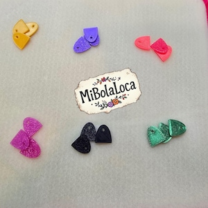May include: A collection of colorful, small, decorative earrings in various shapes and colors, including yellow, purple, pink, black, and green. The earrings are arranged on a light gray surface, with the text "MiBolaLoca" in the center.