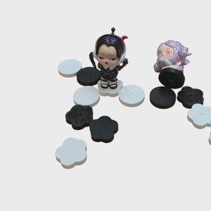 May include: A collection of small figurines and decorative pieces. The figurines include a character in a black and white outfit and another in a purple outfit. Various black, white, and gray circular and flower-shaped pieces are scattered around the figurines.
