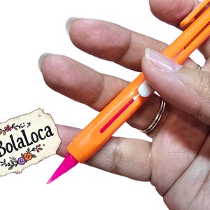 Könnte beinhalten: Ein orangefarbener Mehrfarbenstift mit einer pinkfarbenen Spitze wird in einer Hand gehalten. Der Stift hat einen roten Streifen und weiße Akzente. Ein kleines, dekoratives Etikett mit dem Text "MiBolaLoca" befindet sich in der unteren linken Ecke.