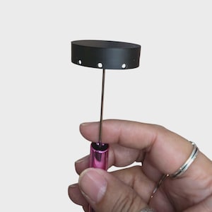May include: A black, oval-shaped jewelry display stand with a pink handle. The stand has a thin metal rod and a black top with three small holes. The stand is designed to hold jewelry.