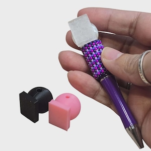 May include: A purple pen with a beaded grip in shades of purple, pink, and white, topped with a clear, square-shaped crystal. Two additional pen toppers, one black and one pink, are also visible.