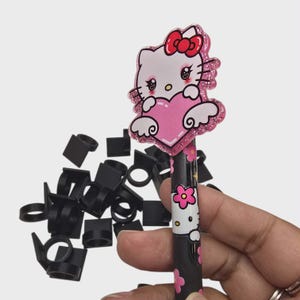 May include: A black and pink Hello Kitty pen with a pink heart and bow design. The pen has a glittery border and is decorated with pink flowers. A pile of small black plastic clips are in the background.