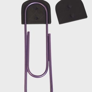 May include: A large, purple paper clip with a black, curved top and a separate black piece. The paper clip is a vibrant purple color and has a smooth, metallic finish. The black pieces have a textured appearance.