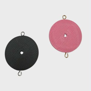 May include: Two round, textured coasters. One is black, the other is pink. Each has a small hole in the center and a metal loop attached at the top and bottom. The coasters are designed to protect surfaces from heat and moisture.