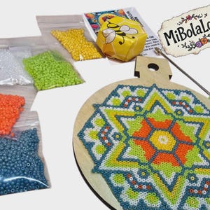 May include: A craft kit with a wooden base featuring a colorful geometric design made of small beads. The kit includes bags of beads in various colors, a tool, and a small box with a bee graphic. The MiBolaLoca logo is visible.