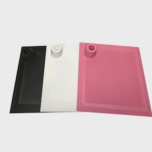 May include: Three square trays in black, white, and pink. Each tray has a raised rim and a circular holder on one corner. The trays are arranged side by side, showcasing their different colors and designs.