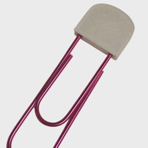 Puede incluir: Un clip de papel grande, de color magenta, con una parte superior redondeada de color blanquecino. El clip de papel metálico tiene un acabado liso y brillante. La parte superior tiene forma rectangular con bordes redondeados. Diseñado para sujetar papeles.