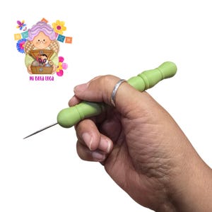 May include: A green needle tool with a rounded handle is held in a hand. The tool has a long, thin, silver needle extending from one end. The handle is a light green color. The image also includes a cartoon illustration with the text "Mi Bola Loca".