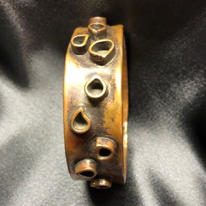 Könnte beinhalten: Ein goldfarbenes Metallarmband mit einer Reihe von tropfenförmigen Ausschnitten. Das Armband ist glatt und hat eine leicht abgenutzte Oberfläche.