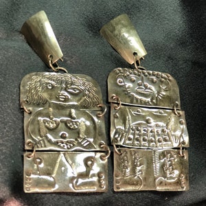 Könnte beinhalten: Ein Paar silberne Ohrringe mit einem dreidimensionalen Design aus stilisierten Figuren. Jeder Ohrring besteht aus drei miteinander verbundenen Teilen, die jeweils ein anderes Design aufweisen. Die Figuren sind in einem einfachen, fast cartoonartigen Stil dargestellt.
