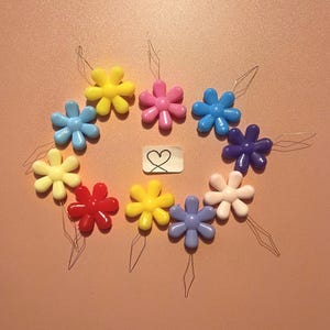 May include: A collection of ten colorful plastic flower-shaped needle threaders arranged in a circle. Each flower features six rounded petals in shades of yellow, pink, blue, purple, red, and white, with a thin wire loop. A small white tag with a hand-drawn heart is in the center.